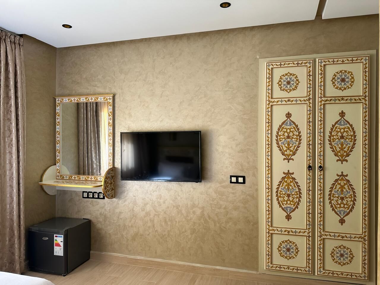 A hotel room with a flat-screen TV mounted on the wall, a small cabinet below it, and a wardrobe with intricate designs.
