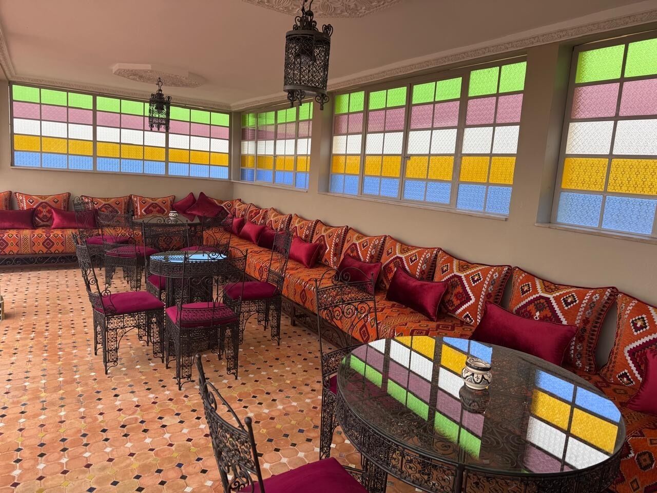 Una habitación con piso estampado, un sofá largo con cojines estampados, una mesa redonda con sillas y ventanas grandes con vidrios de colores.