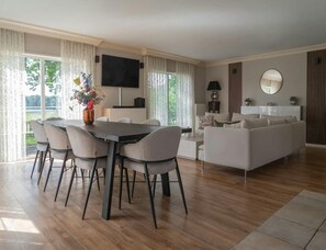 A modern living room with a dining area, a flat-screen TV mounted on the wall, and a large window with curtains. - Chalet With Stunning Lake Vistas (Heinkenszand)