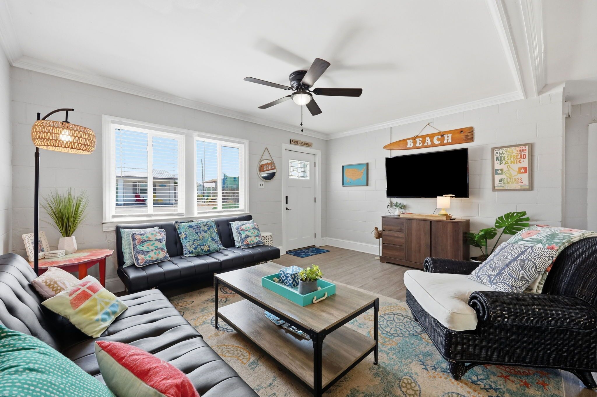 A living room with a black leather sofa, a coffee table, a wicker chair, a ceiling fan, a TV, and a sign that reads 'BEACH'.