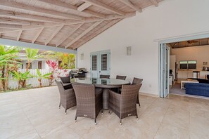 Eine Terrasse im Freien mit einem runden Tisch, Korbstühlen, einem Grill und Blick auf ein Haus mit weißem Zaun und tropischen Pflanzen.
