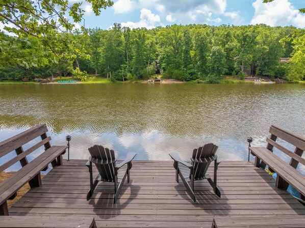 Een houten terras met twee Adirondack stoelen gericht op een kalm meer, omgeven door dichte groene bomen.