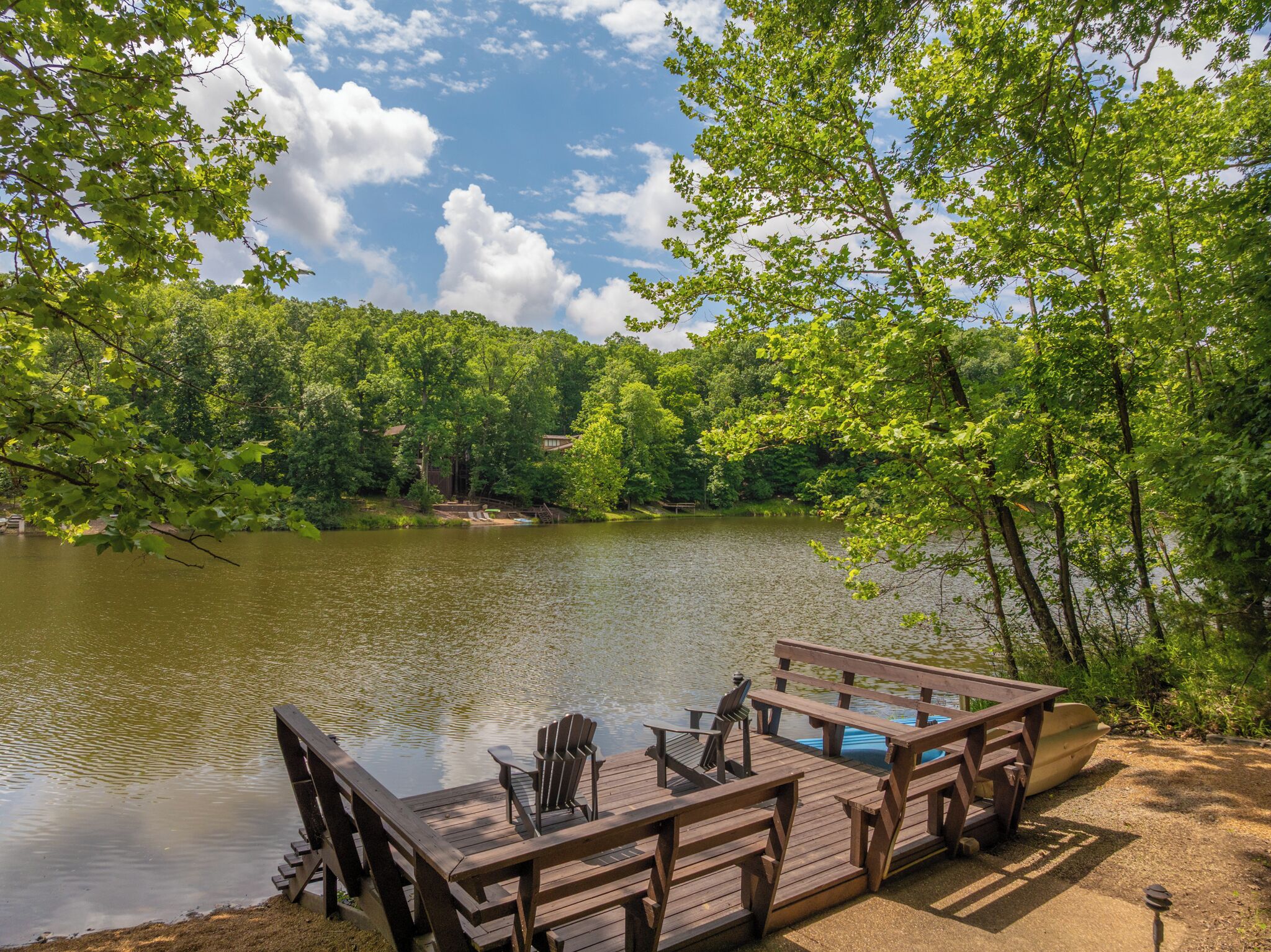 A wooden dock with chairs and a table by a calm lake, surrounded by dense green trees.
