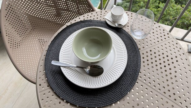 A table with a cup, saucer, and spoon, a glass of water, and a woven placemat. - GA 22 Ape com AR prox. Allianz e A (São Paulo)