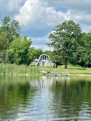 Une maison au bord d’un lac, avec un quai, entourée d’arbres.