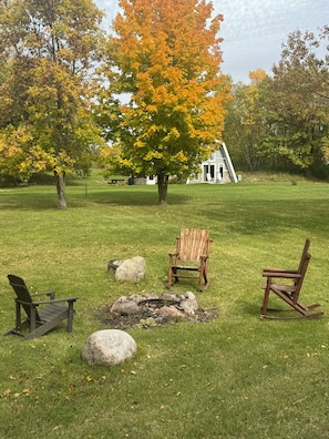 Un espace extérieur verdoyant avec un foyer, deux fauteuils Adirondack et un bâtiment blanc en arrière-plan.