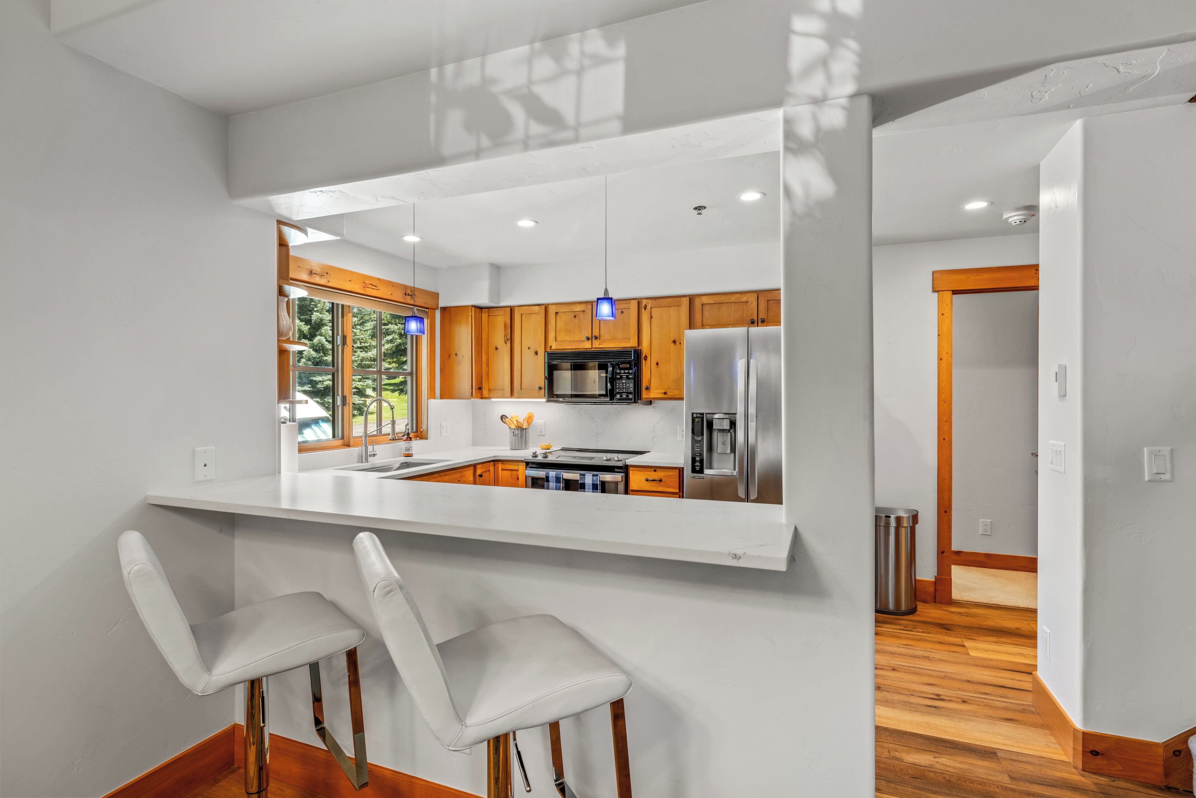 Una cocina moderna con encimeras blancas, armarios de madera, electrodomésticos de acero inoxidable y una barra con taburetes blancos.
