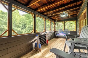 Um deck de madeira com vista para árvores, um ventilador de teto e uma área de estar com mesa e cadeiras.