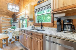 A kitchen with wooden cabinets, granite countertops, a stainless steel dishwasher, a coffee maker, and a window with a view of greenery. - PET Friendly - 2 King Beds -Cedar Brooke Haven (Sevierville)