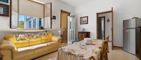 A dining area with a table set for two, a sofa, a fan, and a refrigerator.
