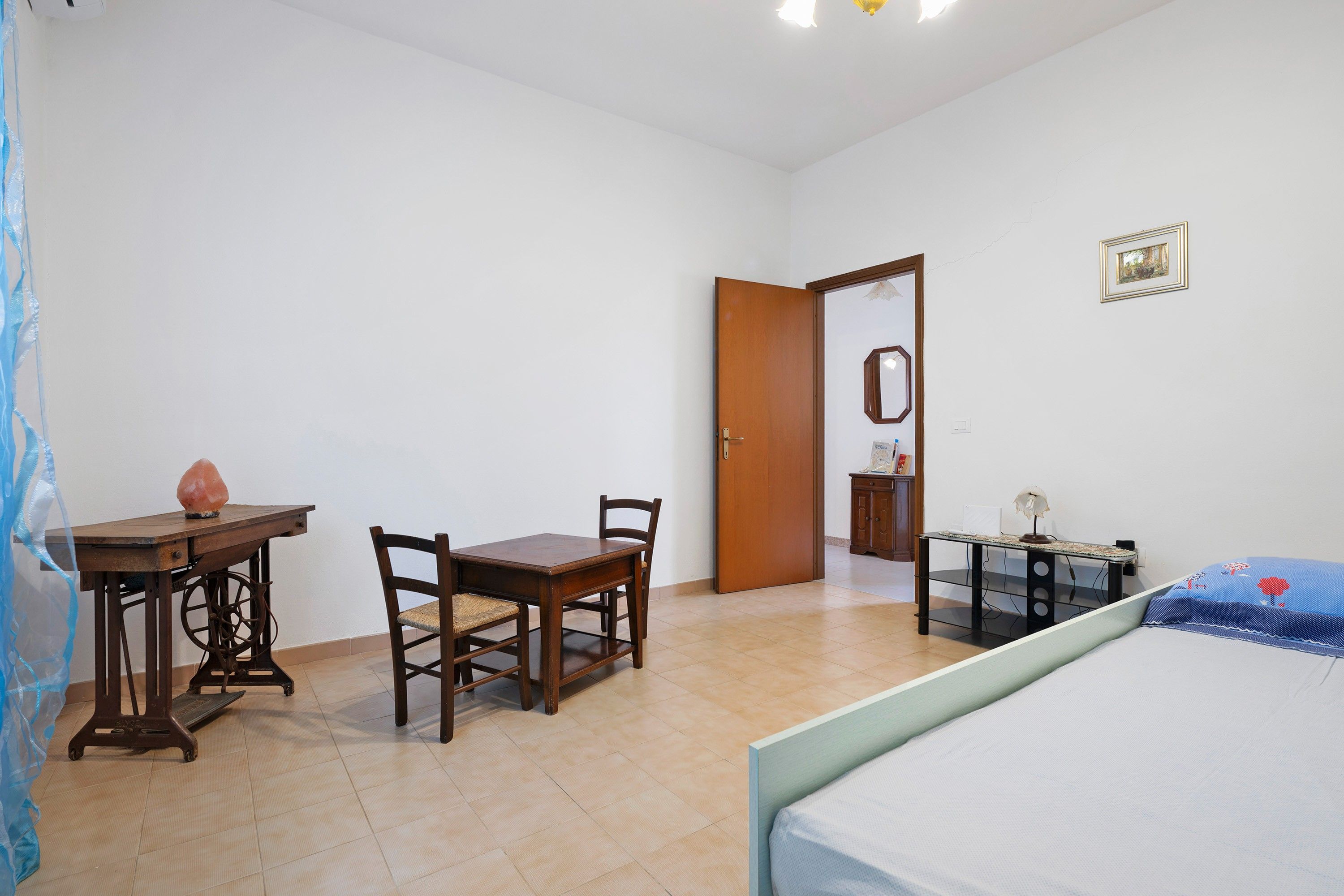 A bedroom with a bed, a wooden table with chairs, a desk with a lamp, and a door leading to another room.
