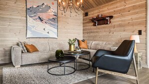 Ein gemütliches Zimmer mit einer Couch, einem Sessel, einem kleinen Tisch, einer Chandelierleuchtre und einem an der Wand befestigten Regal mit Dekorationen.