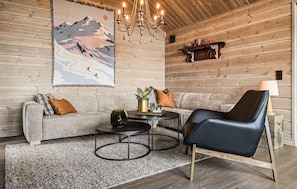 Ein gemütliches Zimmer mit einer Couch, einem Sessel, einem kleinen Tisch, einer Chandelierleuchtre und einem an der Wand befestigten Regal mit Dekorationen.