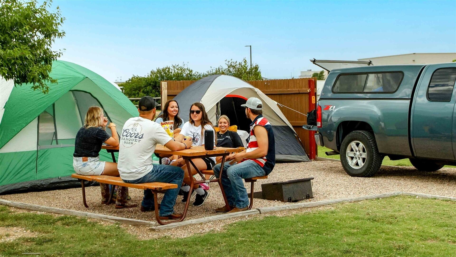 A group of people sitting at a picnic table outdoors with tents and a vehicle nearby.