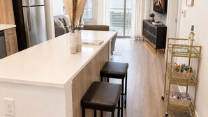 Eine moderne Küche mit einer zentralen Kochinsel, Barhocker, einem Essbereich mit einer Couch, einem Fernseher und einer Zierpflanze.
