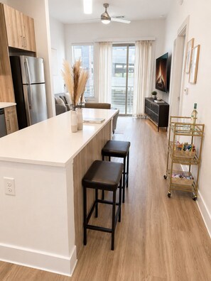 A modern kitchen with a central island, bar stools, a dining area with a sofa, a TV, and a decorative plant. - Riverwalk Oasis (San Antonio)