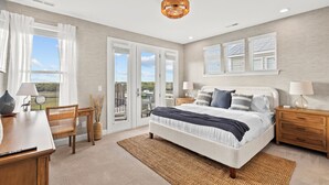 A bedroom with a large bed, a desk, a chair, a lamp, and a view through the window. - Sunsets, Salty Air, and Waterfront Views (Surf City)
