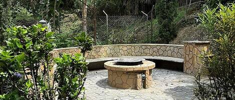 A stone fire pit surrounded by a stone wall, with a grassy area and trees in the background.