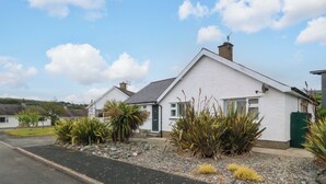 A white house with a sloped roof, a visible chimney, and a small garden with various plants. - 5 Ger Afon (Pwllheli)