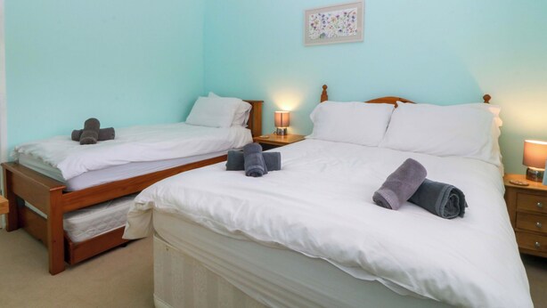 A bedroom with two beds, a bedside table with a lamp, and a wall-mounted light fixture. - 5 Ger Afon (Pwllheli)