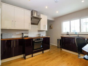 A kitchen with wooden cabinets, a stainless steel range hood, a built-in oven, and a window with blinds. - 5 Ger Afon (Pwllheli)