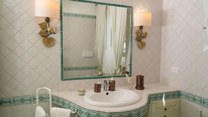 Une salle de bain avec un lavabo blanc, un miroir doté d’un cadre vert et deux lampes murales ornées de motifs de feuilles.