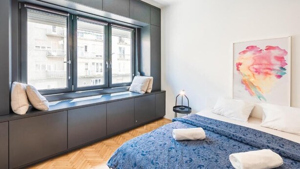 Une chambre à coucher moderne avec une grande fenêtre, un lit avec une literie bleue, une table de chevet avec une lampe et une peinture abstraite aux couleurs vives accrochée au mur.