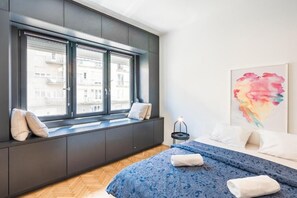 A modern bedroom with a large window, a bed with blue bedding, a bedside table with a lamp, and a colorful abstract painting on the wall. - Bright Studio Parliament Free Public Parking near (Budapest)