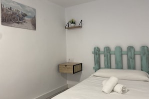 A bedroom with a bed, a nightstand, a wall-mounted shelf with a plant, and a painting of a beach scene. - Casa Gatu 307A by R2R (Cudillero)