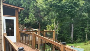 Une terrasse en bois avec des garde-corps, une porte et une vue sur une cour verdoyante et luxuriante, parsemée d’arbres.