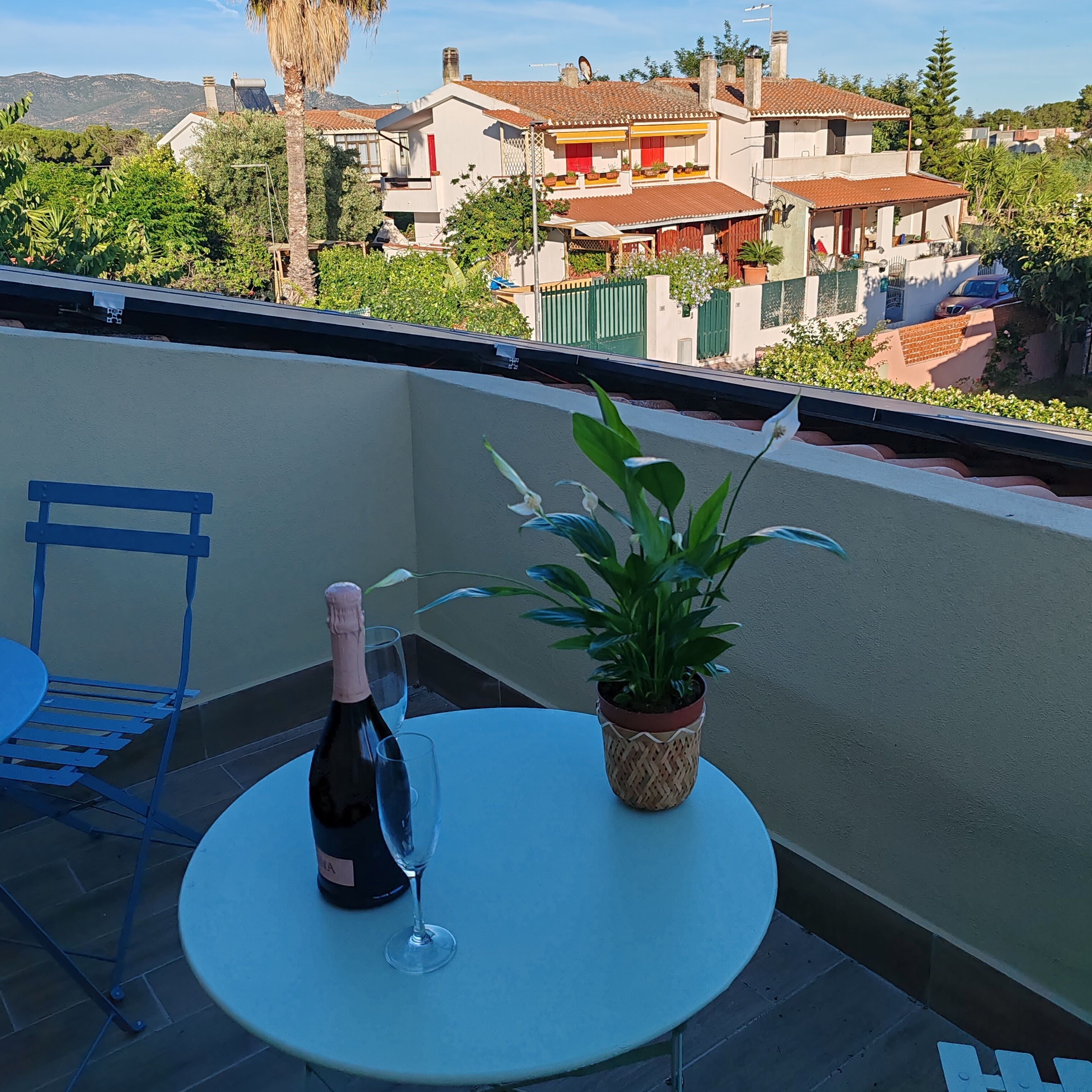 A balcony with a table, a bottle of wine, and a glass, surrounded by greenery and a view of buildings.