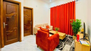 A room with a wooden door, a sofa, a small table, a plant, and a desk with a computer. - Furnished 1-bedroom apartment with a living room, fully equipped kitchen, and Wi-Fi (Brazzaville)