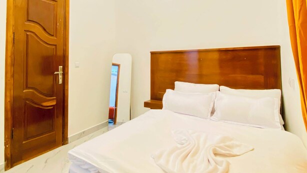 A bedroom with a bed, wooden headboard, white bedding, a ceiling light, an air conditioning unit, and a wooden door. - Furnished 1-bedroom apartment with a living room, fully equipped kitchen, and Wi-Fi (Brazzaville)
