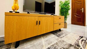 A wooden TV stand with a flat-screen TV, a small statue, and a potted plant. - Furnished 1-bedroom apartment with a living room, fully equipped kitchen, and Wi-Fi (Brazzaville)