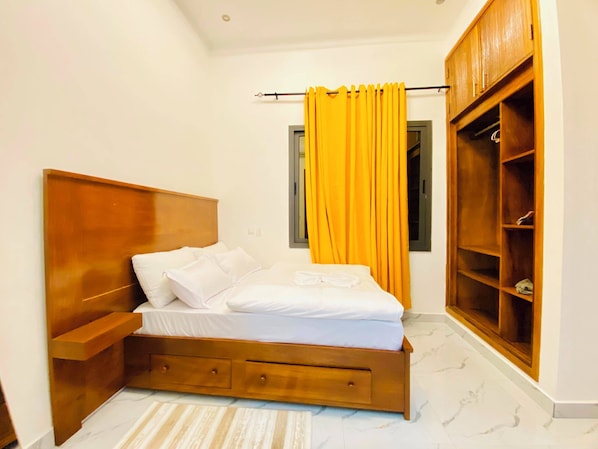 A bedroom with a wooden bed, a small bedside table, a wardrobe, and a window with yellow curtains.