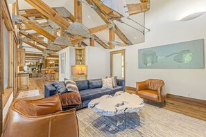 Ein geräumiges Wohnzimmer mit hoher Decke, freiliegenden Holzbalken, einem großen abstrakten Gemälde, einem blauen Sofa, Ledersesseln und einem Couchtisch.