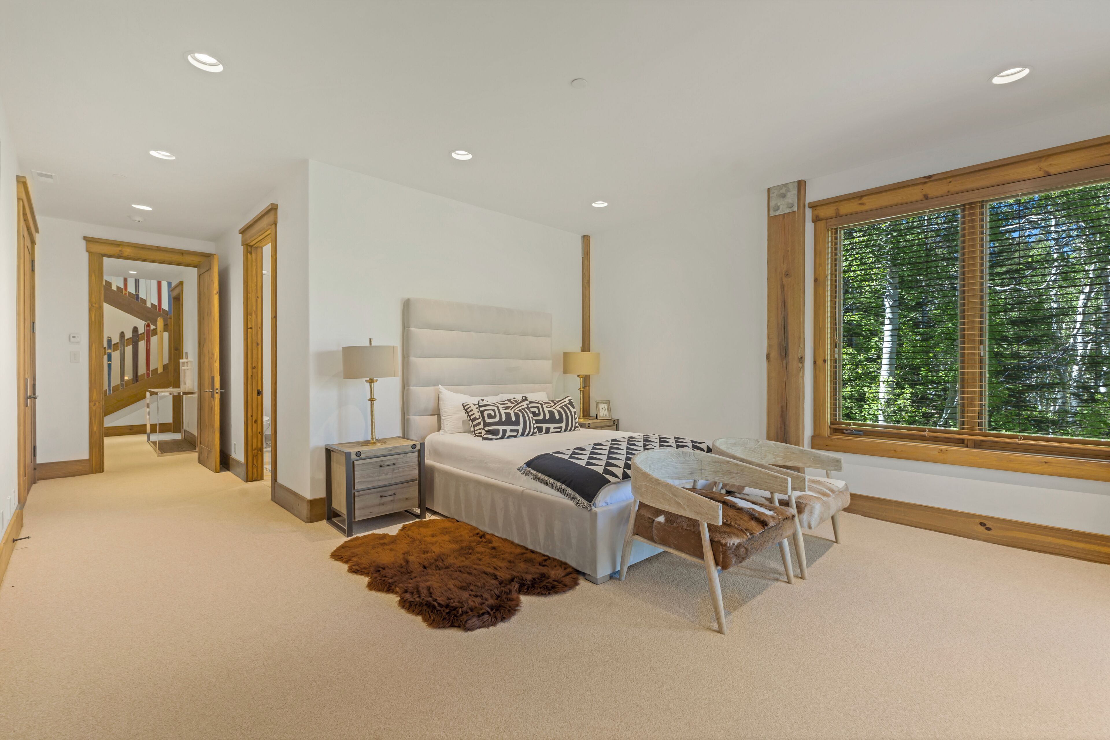 A bedroom with a bed, bedside tables, a lamp, a rug, a window with a view of trees, and a staircase.