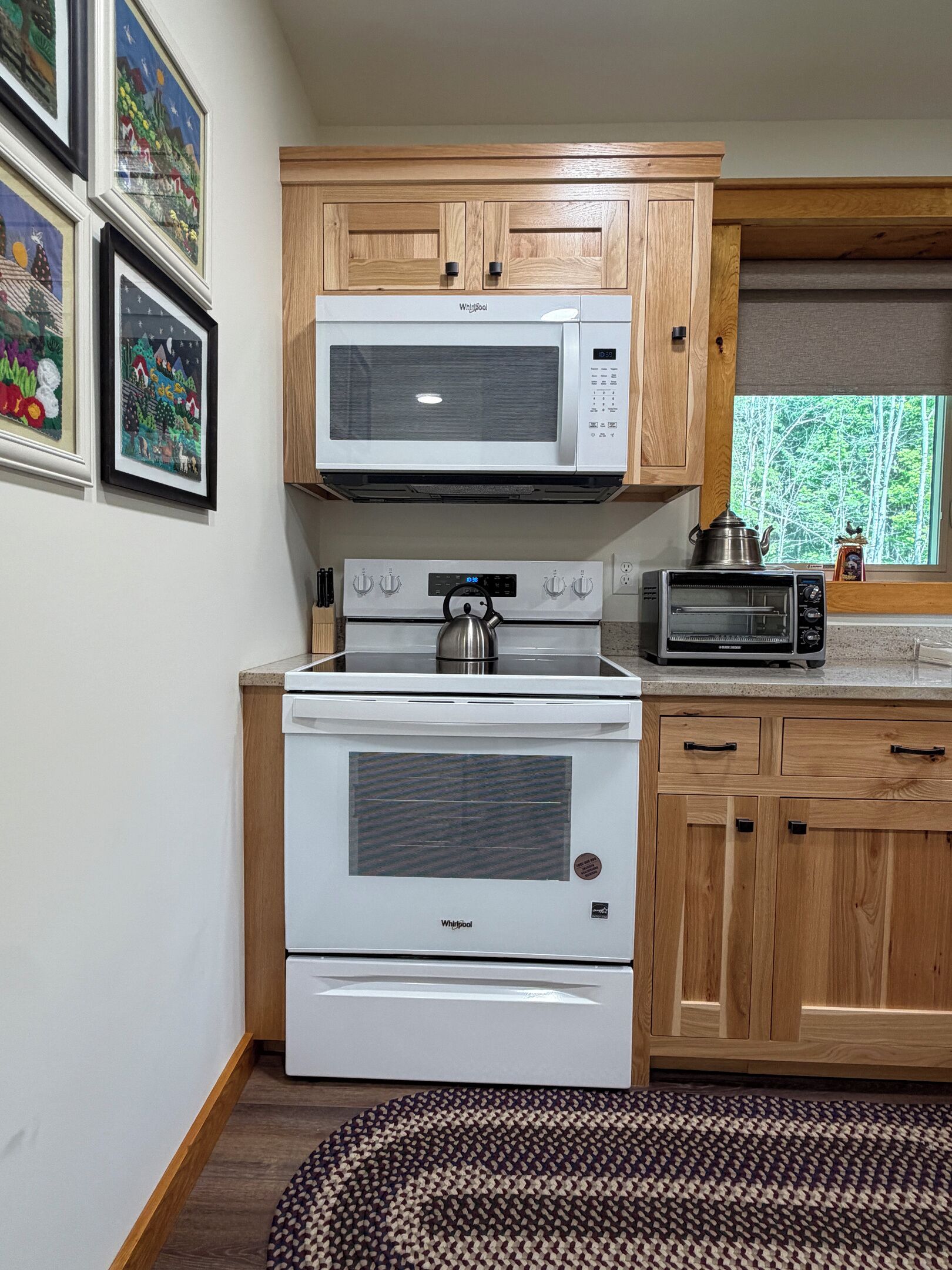 A kitchen with a microwave, stove, and toaster oven. There are wooden cabinets and a window with a view of trees.