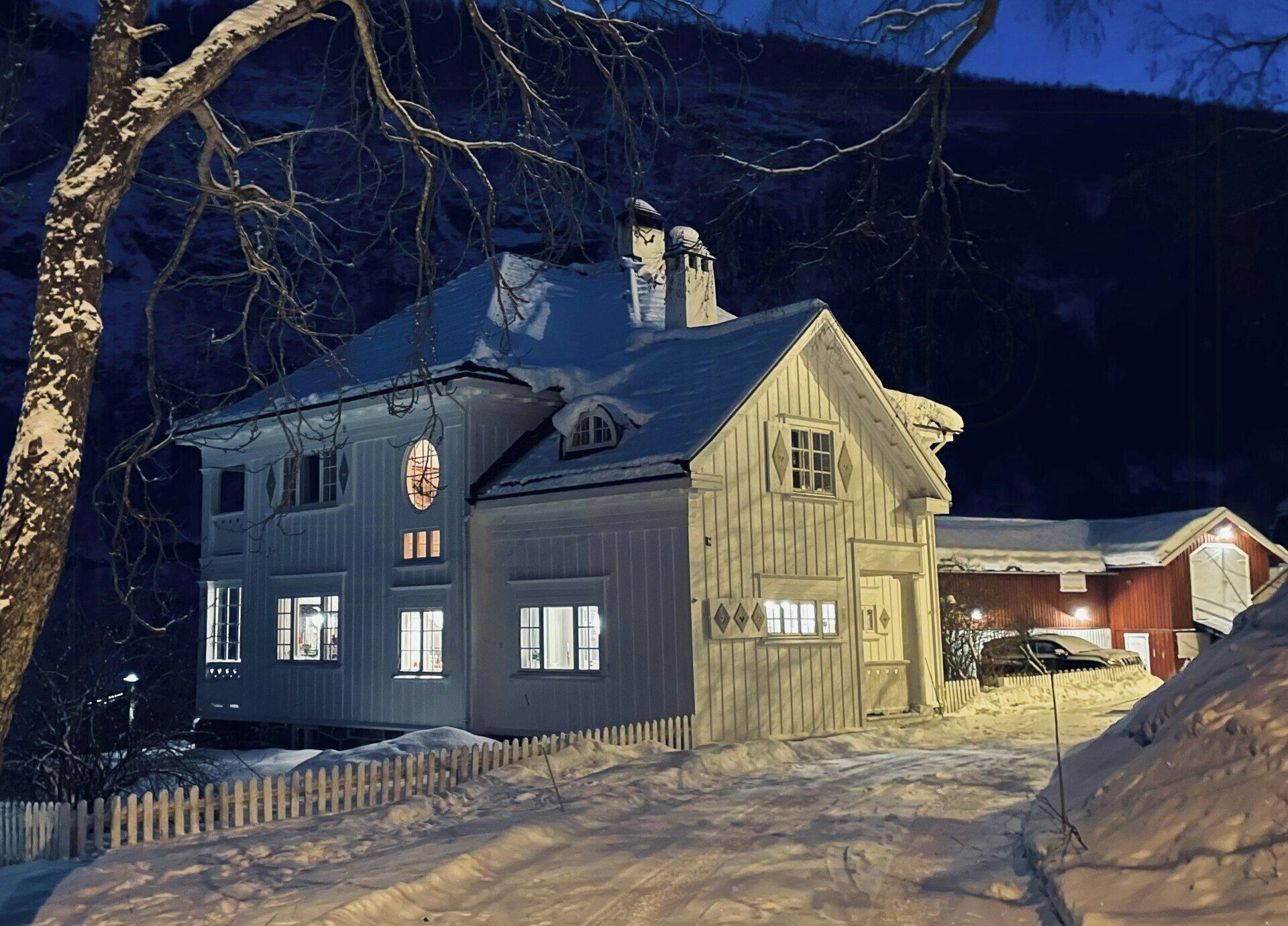 Ett tvåvåningshus byggt av trä, med ett snötäckt tak, ett staket och en synlig skorsten.