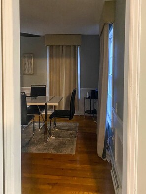 A room with a dining table, chairs, a computer, and a window with curtains.