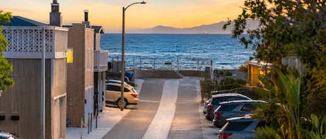 A coastal street with parked cars, a clear view of the sea, and a distant mountain range.