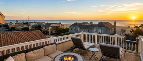 Une terrasse avec un canapé, des chaises et un foyer, offrant une vue sur la plage et l’océan au coucher du soleil.