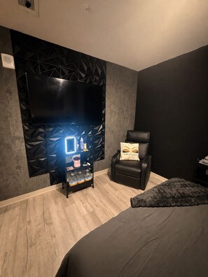 A modern room with a large flat-screen TV mounted on a geometric-patterned wall, a leather chair, a small shelf with toiletries, and a dark-colored bed. - Cozy 2-bedroom High-Rise apt in Downtown Atlanta w/pool, gym, & rooftop access. (Atlanta)