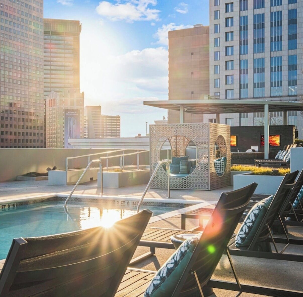 A rooftop pool area with lounge chairs, a seating area with a view of the city skyline, and a pool with a sunlit water surface.