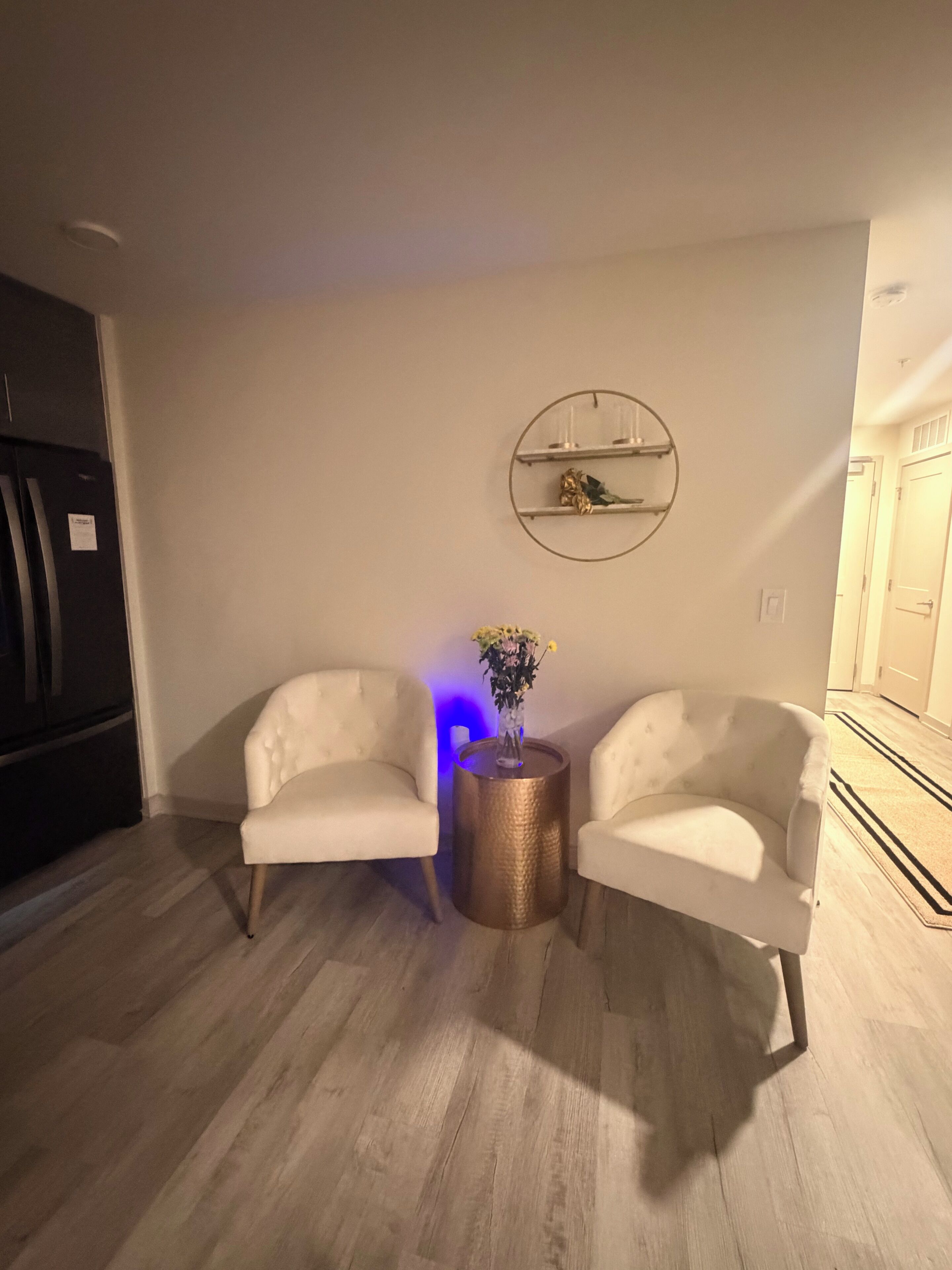 A modern living room with two white armchairs, a round side table with a vase, and a wall-mounted shelf with decorative items.