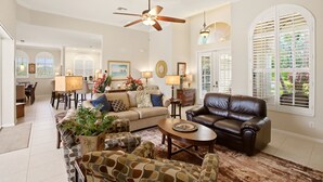 A spacious living room with a ceiling fan, a brown leather sofa, patterned armchairs, and a dining area with a table and chairs. - Upscale 4-bedroom house in peaceful Naples with cool AC, Pool, and Spa (Naples)