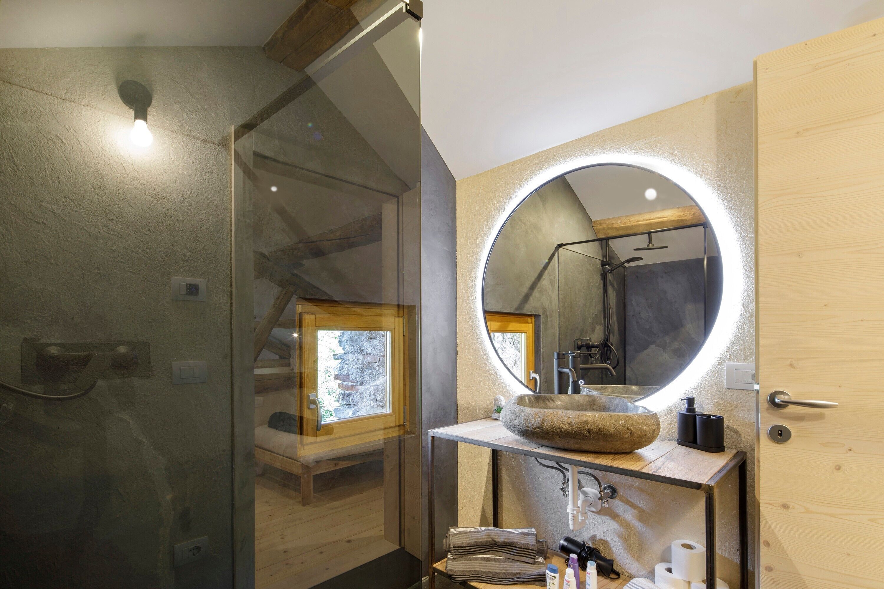 Un bagno moderno con cabina doccia in vetro, mobile lavabo in legno e lavandino in pietra, oltre a un grande specchio con illuminazione integrata.