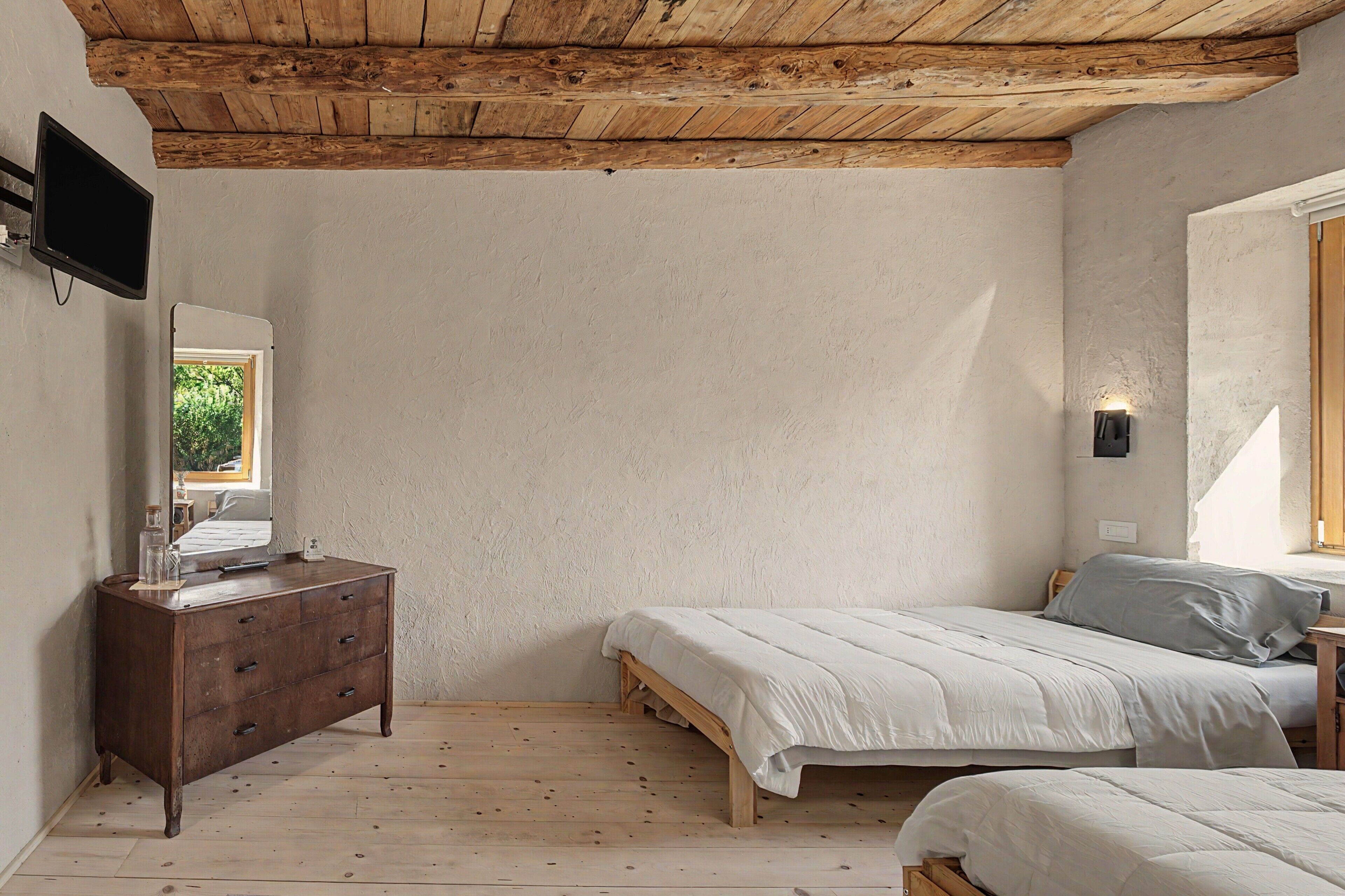 A bedroom with a wooden ceiling, a bed, a dresser, a television, and a window with a view of greenery.