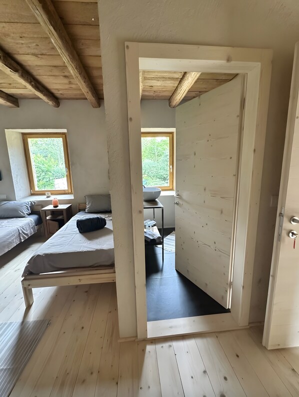 Una habitación con techo de madera, cama con almohada, una pequeña mesa con lámpara y ventana con vista a follaje.