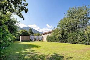 A grassy outdoor area with a stone building, trees, and mountains in the background.